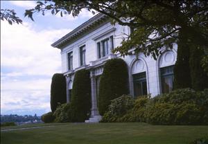 A large white house with arched windows and landscaped yard looks out over a cloudy sky. 